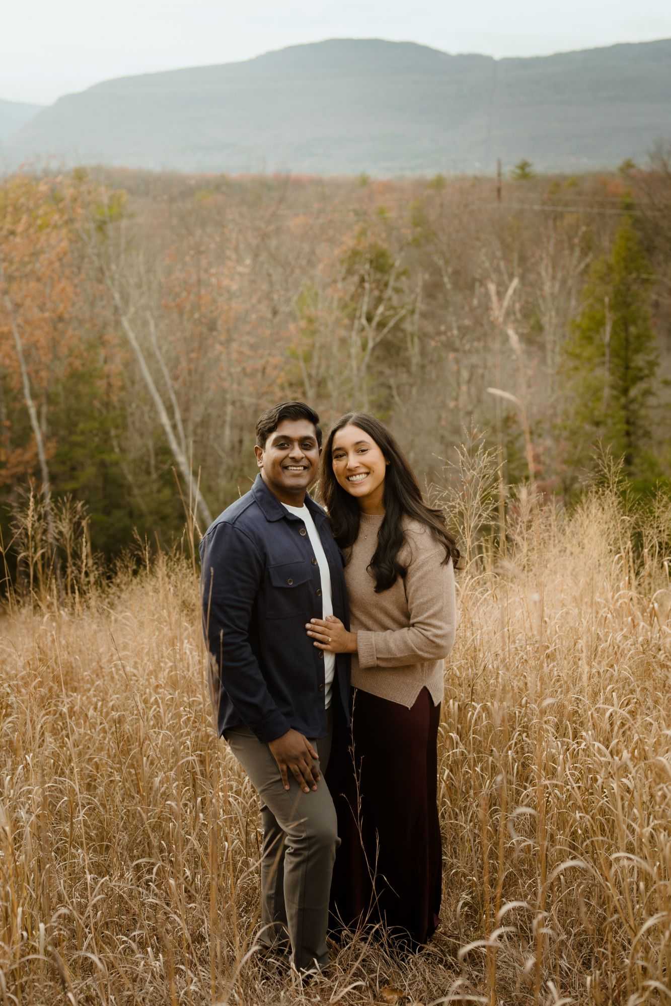 Myra and Rakesh standing together in tall golden grass, smiling at the camera, with autumn hills behind them