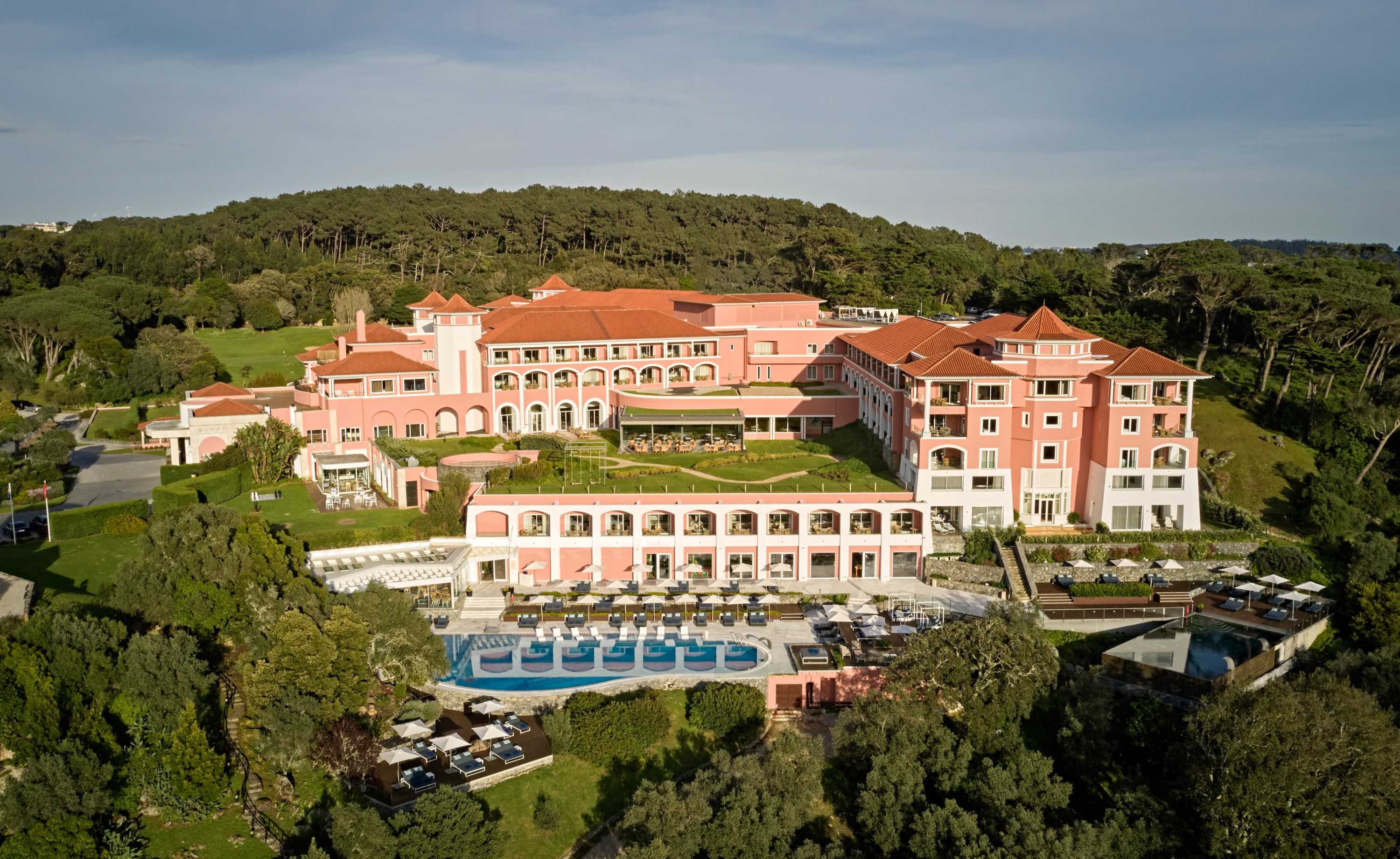 Penha Longa Resort from the air: the pink hotel, terraces and pool in a wooded valley, with green hills behind